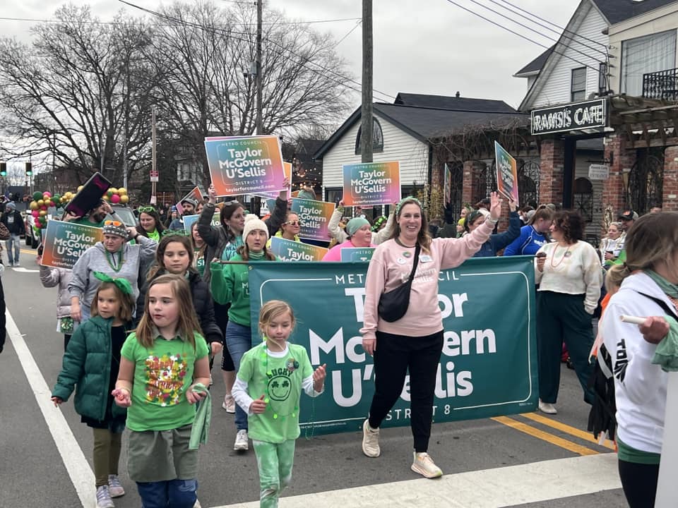 Image depicts the Taylor McGovern U'Sellis campaign marching down a Louisville street during an annual St. Patrick's Day parade. Participants are waving and holding campaign signs with U'Sellis herself at the head.
